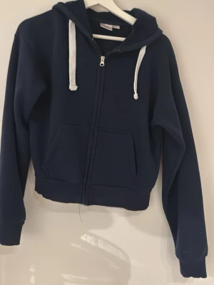 Mörkblå hoodie med dragkedja - Mörkblå hoodie med vit dragsko och dragkedja framtill. Hoodien har två stora fickor på magen och ribbade muddar vid ärmslut och nederkant. Perfekt för en avslappnad stil och enkel att matcha med jeans eller mjukisbyxor.