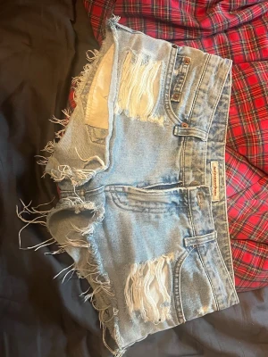 ljusblå slitna jeansshorts - Ljusblå jeansshorts från Hot Pepper med låg midja, råa fransiga kanter och slitna detaljer både fram och bak. Klassisk femficksmodell med knappgylf och Hot Peppers-lapp bak. Perfekta för en avslappnad och trendig sommarstil.