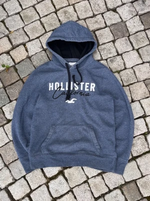 Blå Hollister Hoodie - Snygg blå hoodie från Hollister med stor vit och svart logga på bröstet där det står 'Hollister California'. Hoodien har en klassisk känguruficka framtill och justerbar huva med svarta snören. Mjuk insida och avslappnad passform, perfekt för chill dagar.