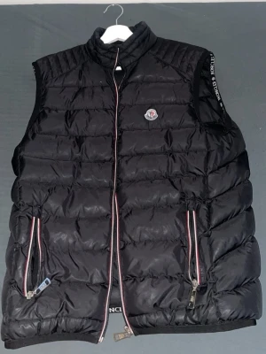 Svart dunväst från Moncler - Snygg svart dunväst från Moncler med hög krage och två fickor med dragkedja. Västen har Moncler-logga på bröstet och detaljer i rött, vitt och blått längs dragkedjorna. Perfekt för lager-på-lager och streetwear-stil.