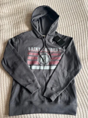 Grå Saint Laurent hoodie  - Riktigt fet hoodie från Saint Laurent, hoodien är stilren och passar riktigt bra. Tyvärr måste jag sälja hoodien eftersom att den inte passar mig längre. Hoodien är grå och i storlek S. Tveka inte på att ställa frågor!🤙