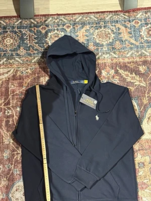 Marinblå hoodie från Polo Ralph Lauren - Snygg marinblå hoodie från Polo Ralph Lauren med dragkedja framtill och klassisk huva. Ikoniska loggan i vitt på bröstet. Tillverkad i mjukt bomullsmaterial och har ribbade muddar vid ärmslut och nederkant. Perfekt för en avslappnad och stilren look.