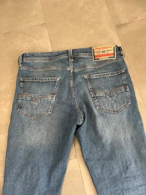Blå raka jeans från Diesel - Säljer ett par klassiska blå jeans från Diesel med raka ben och snygg slitning. Jeansen har fem fickor, tydliga sömmar och en patch med Diesel-logga bak i midjan. Perfekta för en avslappnad och trendig look.