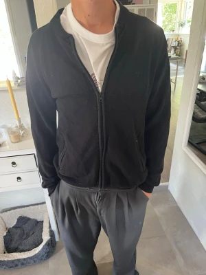 Svart ziptröja med ribbade detaljer - Säljer en svart ziptröja med ribbade muddar och krage. Tröjan har två fickor med dragkedja och är tillverkad i ett mjukt material. Enkel och stilren design som passar till det mesta. Perfekt för dig som gillar en clean look.