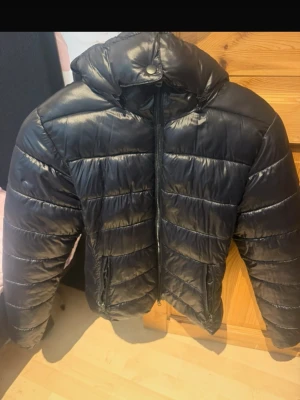 Svart glansig pufferjacka med huva  - Säljer en svart pufferjacka med glansig finish och huva. Jackan har dragkedja framtill, två sidofickor med dragkedja och breda quiltade paneler. Perfekt för kalla vinterdagar och ger en cool streetstyle-look. Använd en säsong