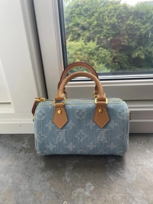 Blå denim handväska med monogram - Snygg liten handväska i ljusblå denim med klassiskt monogrammönster och detaljer i brunt läder. Väskan har två korta handtag och guldfärgade metalldetaljer. Perfekt accessoar för att lyfta din outfit. band finns 