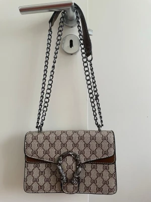 Beige axelväska med kedjerem - Snygg beige axelväska med monogrammönster och dubbla kedjeremmar i metall. Väskan har ett dekorativt spänne framtill i form av två ormar och detaljer i brunt läder. Perfekt storlek för det viktigaste och ger en lyxig känsla till din outfit.