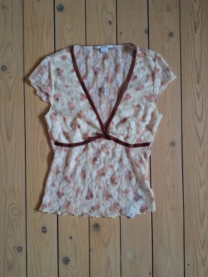 Söt Kimchi Blue Spetstopp - Söt blommig spetsblus i ljusbeige med rosa blommor och bruna sammetsband längs v-ringningen. Toppen täcker bröstet men är transparent på mage och rygg. Det står storlek M men passar XS/S. Pagget är elastiskt. Längd ca 47 cm.