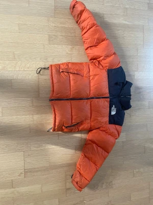 Orange pufferjacka från The North Face - Säljer en fet orange pufferjacka från The North Face med svart axelparti och huva. Jackan har två sidofickor med dragkedja,dragkedjan går inte att dra hela vägen ner,  justerbar nederkant och stora puffiga sektioner. Perfekt för kalla vinterdagar och riktigt snygg streetstil. Skriv vid frågor och prisförslag 