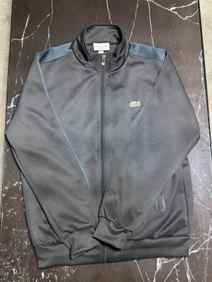 Svart ziptröja från Lacoste - Snygg svart ziptröja från Lacoste med diskret blå detalj längs axlar och bröst. Tröjan har hög krage, dragkedja framtill och ribbade muddar. Klassisk Lacoste-logga i grönt och vitt på bröstet. Perfekt för en sportig och clean look.