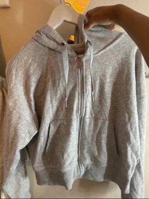 Grå zip hoodie från Calvin Klein - Snygg grå zip hoodie från Calvin Klein med huva och dragsko. Tillverkad i mjukt bomullsmaterial och har två fickor framtill. Perfekt för en chill och avslappnad stil. Dragkedjan har Calvin Klein-logga för extra detalj.
