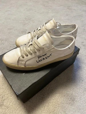 Saint Laurent - Säljer ett par stilrena vita sneakers från Saint Laurent i storlek 43. Skorna har den klassiska Saint Laurent designen med detaljer och logga på sidan. De är använda och har tydliga tecken på användning, bland annat smuts och slitage på sulan och ovandelen, vilket syns på bilderna. Trots det är de fortfarande snygga och går absolut att använda.  Storlek: 43 Märke: Saint Laurent Färg: Vit Skick: Använt skick, se bilder för detaljer Kartong: Medföljer