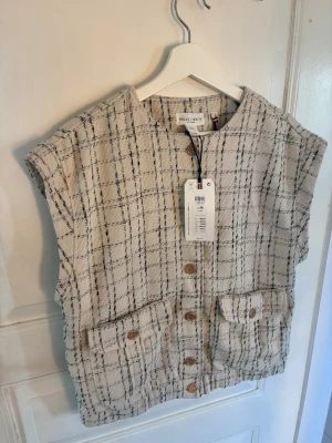 Beige väst från Holly & Whyte - Stilren beige väst från Holly & Whyte med rutigt mönster i svart. Västen har rund halsringning, knappar framtill och två stora fickor. Tillverkad i ett strukturerat tyg som ger en trendig look. Perfekt för att lyfta din outfit.