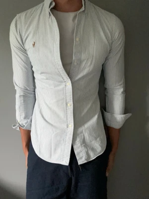 Randig skjorta från Ralph Lauren - Snygg ljusblå och vit randig skjorta från Ralph Lauren med klassisk button-down krage och broderad logga på bröstet. Skjortan har långa ärmar och smal passform, perfekt till jeans eller chinos. Tillverkad i bomull för en skön känsla.