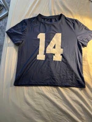 Blå t-shirt med stor siffra 14 - Säljer en blå t-shirt med en stor vit siffra 14 tryckt på framsidan. T-shirten har korta ärmar och en klassisk rund halsringning. Perfekt för dig som gillar sportig stil eller vill sticka ut med en enkel men cool detalj.