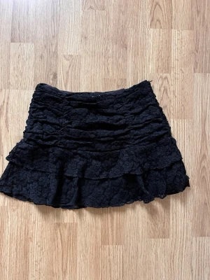 Svart volangkjol i spets - Kort svart kjol med flera lager volanger och vacker spetsdetalj. Kjolen har en feminin och trendig look som passar perfekt till olika stilar. Materialet är mjukt och följsamt för en bekväm känsla.