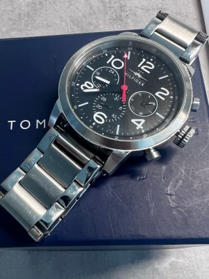 Stilren herr silverklocka från Tommy Hilfiger - Snygg och modern klocka från Tommy Hilfiger med svart urtavla, tydliga vita siffror. Klockan har en robust länk i silverfärgat metall och flera små urtavlor för extra funktioner. Perfekt accessoar för en trendig look. Herr modell. 