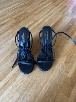 Svarta snörade pumps  - Säljer ett par svarta pumps med tunna remmar och snörning runt ankeln. Skorna har öppen tå och ikoniska detaljer. Slitage finns på bild syns inte vid Andvändning 