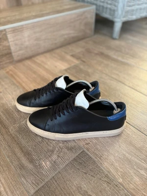 Axel Arigato Clean 90 sneakers  - Axel arigato clean 90 sneakers i läder med vit plös och blå detalj vid hälen. Skorna har svarta skosnören och en vit sula som ger en klassisk look. Fräscht skick storlek 45 