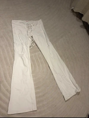 Vita Gina Jeans - Säljer ett par vita bootcut jeans med snygg snörning framtill. Jeansen har låg midja och lätt utsvängda ben. Materialet ser ut att vara bomull med lite stretch för skön passform. Perfekta för dig som vill ha en trendig och unik look.