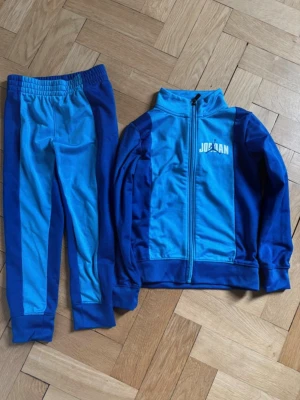Blått Jordan-set - Snyggt träningsset från Jordan i två nyanser av blått. Setet består av en zip-jacka med hög krage och logga på bröstet samt matchande byxor med resår i midjan. Materialet är mjukt och stretchigt, perfekt för sport eller chill. Byxorna är lite noppriga. 
