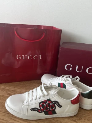 Vita sneakers  - Säljer ett par vita skor  i läder med klassiska gröna och röda ränder på sidan samt en broderad orm på utsidan. Baksidan har metallicröda och gröna detaljer. Snygg och tidlös modell med vita skosnören och låg profil. Skorna är använda 1 gång endast och är i helt nytt skick utöver. Det är inga skador och skorna är som nya