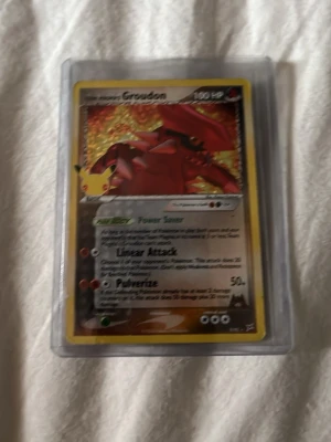 Pokémonkort Groudon holo - Säljer ett Pokémonkort med Groudon-motiv. Kortet är holografiskt med gul ram och har tydliga attacker och effekter. Det ligger i ett genomskinligt plastskydd. Perfekt för samlare eller Pokémonfans. Från 2004