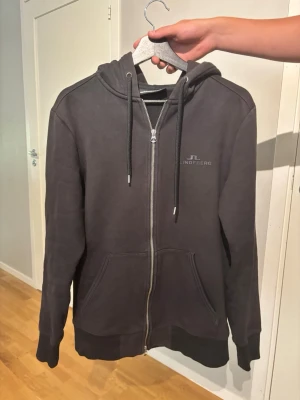 Svart hoodie med dragkedja J.Lindeberg - Säljer en svart hoodie från J.Lindeberg med dragkedja framtill, två fickor och justerbar huva med snören. Diskret logga på bröstet. Tillverkad i mjukt bomullsmaterial som är skönt att ha på sig. Perfekt för en chill och stilren look. Sparsamt använd 