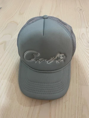 Grå meshkeps med broderad text - Snygg grå keps med böjd skärm och mesh på baksidan för extra ventilation. Framtill finns en broderad text i matchande ton. Justerbar baktill för perfekt passform. Perfekt för en avslappnad streetwear-look.