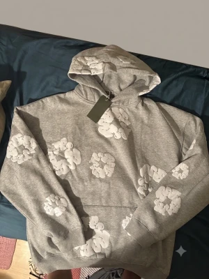 Grå hoodie med vita blommor Denim Tears - Snygg grå hoodie från Denim Tears med vita blommor tryckta över hela tröjan. Klassisk huva och känguruficka framtill. Perfekt för dig som vill sticka ut med en unik och trendig stil.