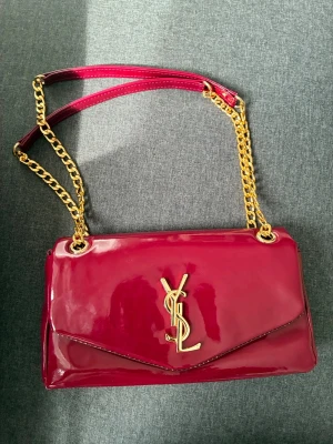 Mörkröd YSL handväska med kedja - Snygg mörkröd handväska från YSL i glansigt lackmaterial. Väskan har guldiga kedjeaxelband och ett stort YSL-emblem framtill. Stilren kuvertform som ger en lyxig känsla och passar perfekt till din outfit.