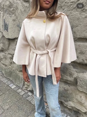 Missäy poncho  - Snygg beige poncho från missäy med vida trekvartsärmar och knytskärp i midjan. Kappan har hög krage och en dekorativ gul knapp upptill. Perfekt för dig som vill ha en stilren och trendig look. Materialet ser ut att vara mjukt och lite tjockare tyg.