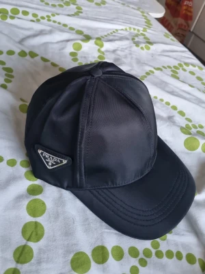 Svart keps (Cordell kedja) Bundle - Snygg svart keps från Prada i glansigt nylonmaterial med klassisk böjd skärm och ikonisk metallogo på sidan. Perfekt accessoar för en trendig och stilren look. Justerbar baktill för bästa passform. DUO man får med cordellkedja (värde 159 kr)