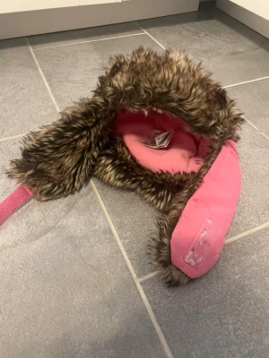 Rosa trappermössa med fuskpäls för barn - Säljer en rosa trappermössa för barn med mjuk fuskpäls i brunt och beige. Utsidan är i rosa tyg och insidan är fodrad med rosa fleece för extra värme. Mössan har öronlappar och kardborreband under hakan.