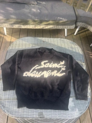Svart stickad Saint Laurent-tröja - Säljer en svart stickad tröja från Saint Laurent med vit logotyp i graffiti-stil på bröstet. Tröjan har rund hals, långa ärmar och ribbstickade muddar vid ärmslut och nederkant. Perfekt för dig som gillar streetwear och exklusiva märken.