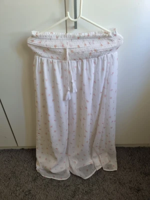 Vit långkjol med blommor från Gina Tricot - Säljer en superfin vit långkjol från Gina Tricot med små rosa blommor och smock i midjan. Kjolen har en dragsko med tofsar och är gjord i ett lätt, luftigt material som passar perfekt till varma dagar.