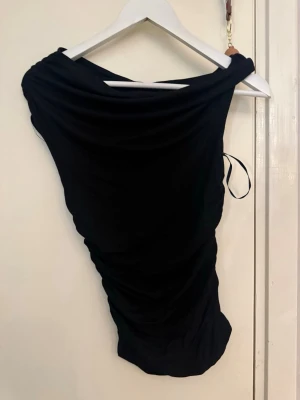 Svart oneshoulder topp - Säljer en svart oneshoulder topp med draperad design och åtsittande passform. Toppen är ärmlös och har en cool, asymmetrisk skärning som ger en trendig look. Perfekt för dig som vill sticka ut lite extra.