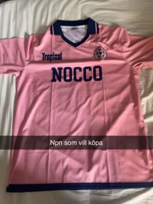 Rosa NOCCO sporttröja med blå detaljer - Säljer en rosa sporttröja med mörkblå detaljer och tryck från NOCCO och Tropical. Tröjan har V-ringad krage med blå kant och korta ärmar. Perfekt för träning eller match.