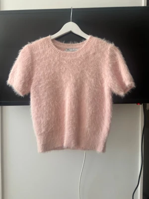 Ljusrosa fluffig stickad tröja Zara - Supersöt ljusrosa stickad tröja från Zara med korta ärmar och fluffig, mjuk textur. Rund halsringning och ribbade kanter ger en clean look. Perfekt för dig som vill ha något gulligt och trendigt i garderoben. Det är en storlek L men är väldigt liten i storleken, skulle säga storlek S