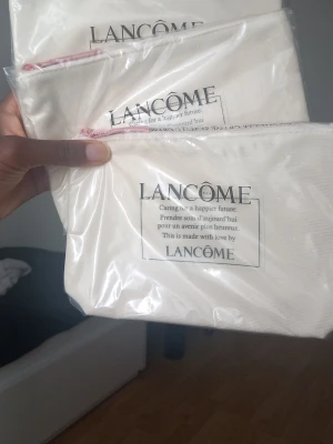 Vita necessärer från Lancôme - Säljer en stilren vit necessär från Lancôme med svart text och dragkedja i rosa. Perfekt för smink eller småprylar. Praktisk storlek och enkel design med tryckt budskap på framsidan. Passar att ha i väskan eller på resan.