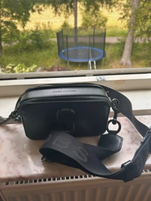 Svart axelväska Marc Jacobs - Snygg svart axelväska från Marc Jacobs med bred justerbar axelrem och präglad logga framtill. Väskan har två dragkedjor upptill och är tillverkad i ett tåligt, strukturerat material. Perfekt för dig som vill ha en stilren och trendig väska.