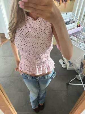 Rosa prickig ärmlös topp med volang - Supersöt rosa topp med små svarta prickar, smockad midja och volangkant nedtill. Toppen är ärmlös och har en figurnära passform som ger en feminin siluett. Perfekt till jeans eller kjol för en trendig look.