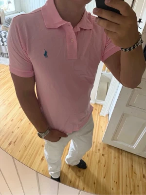 Ralph lauren rosa piké - Säljer en polo Ralph lauren ljusrosa pikétröja med klassisk krage och knappar framtill. Tröjan har korta ärmar och en blå broderad logga på bröstet. Tillverkad i mjuk bomull och har en slim passform som sitter snyggt på kroppen.