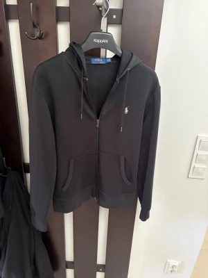 Svart hoodie från Polo Ralph Lauren - Säljer en svart hoodie med dragkedja från Polo Ralph Lauren. Klassisk design med huva, dragsko och två fickor framtill. Liten vit logga broderad på bröstet. Perfekt för en avslappnad och stilren look.