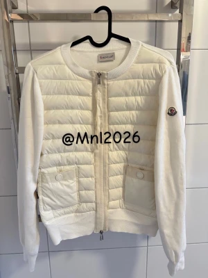 Vit quiltad cardigan från Moncler - Snygg vit cardiganjacka från Moncler med quiltad front och stickade ärmar. Jackan har två fickor med knappar, rund halsringning och dragkedja framtill. Moncler-logga på ärmen ger en exklusiv touch. Perfekt för lager på lager och stilrena outfits.