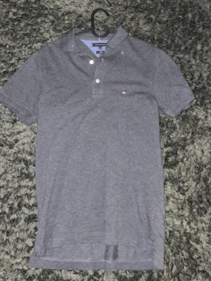 Grå pikétröja från Tommy Hilfiger - Säljer en klassisk grå pikétröja från Tommy Hilfiger med korta ärmar och liten logga på bröstet. Tröjan har krage och knappar framtill, perfekt till både jeans och shorts. Enkel och stilren design som passar till det mesta.