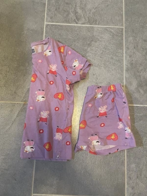 Peppa Pig pyjamas med shorts för barn - Söt lila pyjamas med kortärmad tröja och shorts för barn, dekorerad med Peppa Pig och hjärtan i rött och gult. Perfekt för varma nätter och mysiga kvällar. Mjukt och bekvämt material, troligen bomull. Motiv som passar barn och fans av Peppa Pig.