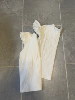 2 Vita toppar med spetsdetaljer för barn - 2 Söta vita toppar för barn i bomull med korta ärmar och spetsdetaljer vid axlarna. Perfekt för en fräsch och enkel look. Toppen har rund halsringning och är lätt att matcha med olika stilar.