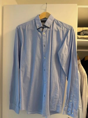 Ljusblå skjorta med småmönster medium - Snygg ljusblå skjorta med diskret småmönster och button-down krage. Skjortan har långa ärmar och vita knappar. Tillverkad i ett tunt, mjukt bomullstyg som känns bekvämt mot huden. Perfekt för dig som gillar stilrena detaljer. Medium