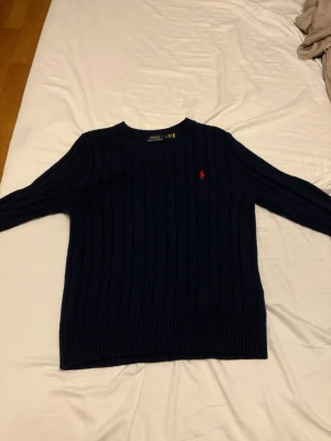 Mörkblå kabelstickad tröja - Mörkblå kabelstickad tröja från Polo Ralph Lauren med rund halsringning och röd broderad logga på bröstet. Tröjan har långa ärmar och ribbade muddar vid ärmslut och nederkant. Klassisk och tidlös design i mjukt material.